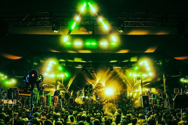 Vibrant night stage at Lost Paradise festival with green laser light show, triangular light rigging, and a dancing crowd holding glowing totems.